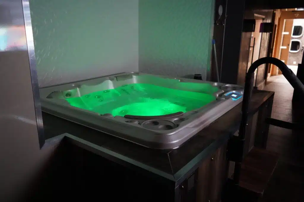 Un bain à remous éclairé par une lumière verte, entouré de bois foncé et de métal, crée une ambiance relaxante dans une pièce moderne et faiblement éclairée.